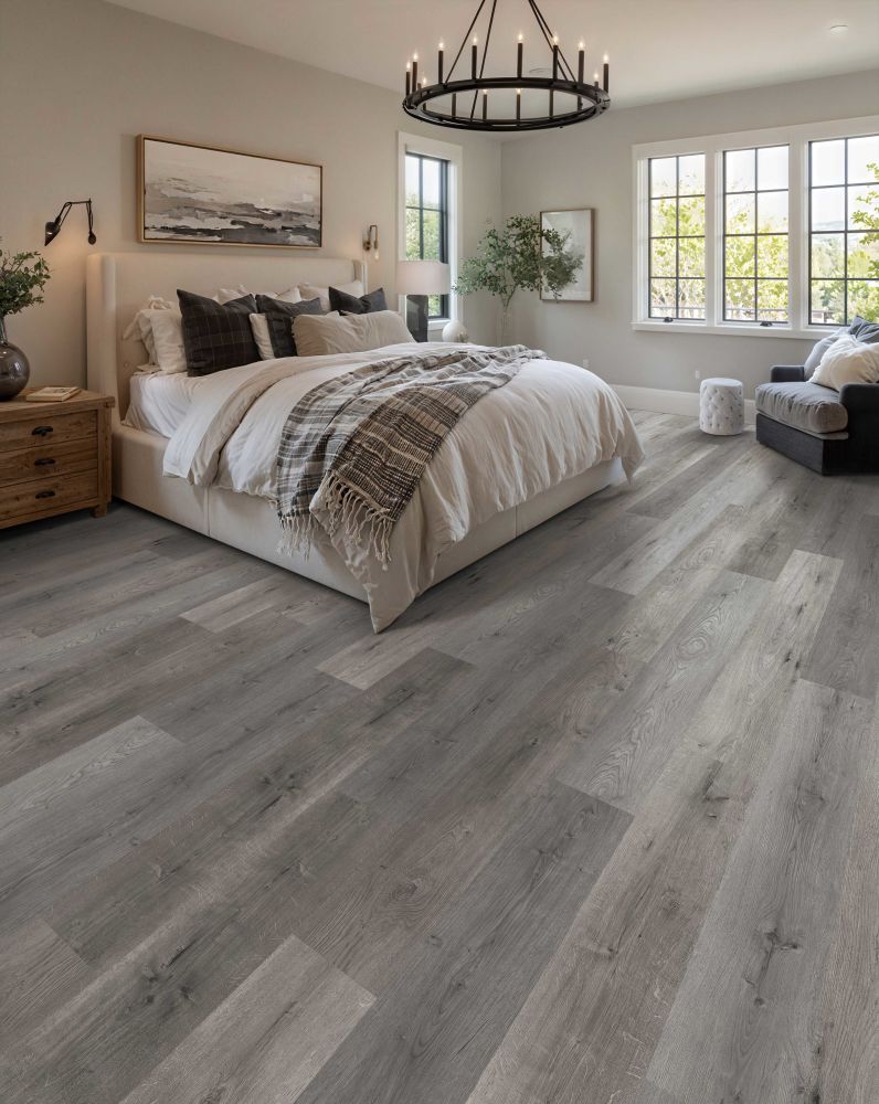 Sunlit master bedroom with a beige upholstered bed, white bedding, plaid throw, and gray wood-plank flooring.