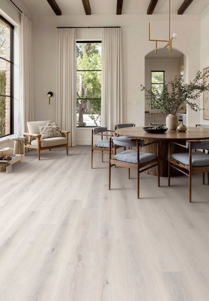 Bright open dining area with a round wooden table and six blue-gray cushioned chairs, large windows with white curtains, and light wood floors.
