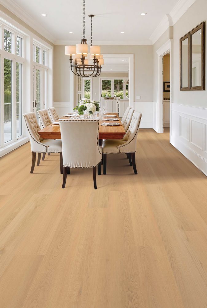 Bright dining room with a wooden table set for six, beige upholstered chairs, a chandelier, and large windows overlooking greenery.