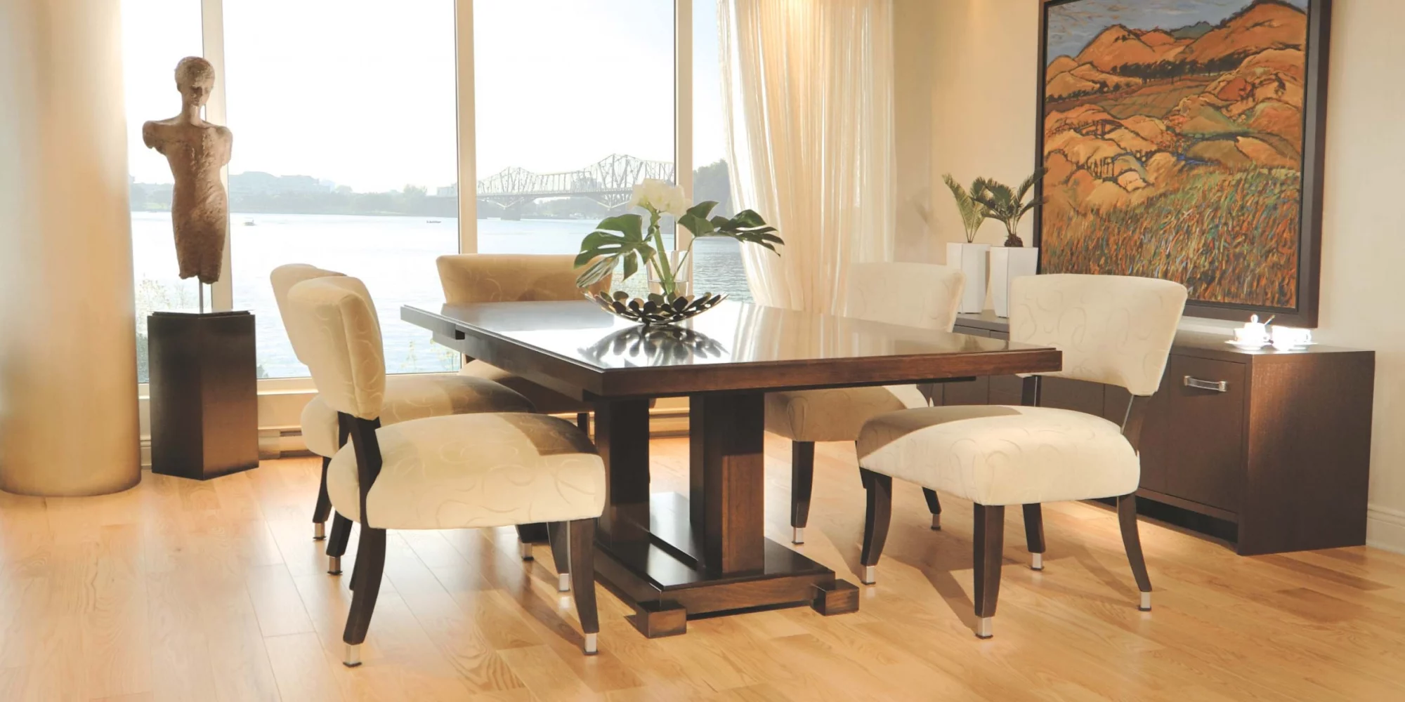 Modern dining room with a glass-top table, six cream upholstered chairs, a sculpture on a pedestal, and a river view outside the window.